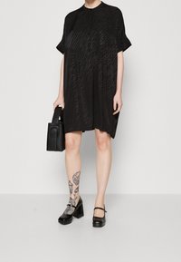 Abito nero a maniche corte con un motivo a onde testurizzate, di lunghezza al ginocchio, con chiusura a bottoni, indossato con una piccola borsa nera e scarpe con tacco grosso.