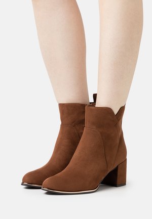 Braune Wildleder-Stiefeletten mit Blockabsätzen, die an nackten Beinen vor einem weißen Hintergrund getragen werden.