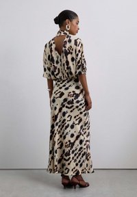 Una mujer que lleva un vestido largo de leopardo beige y negro con un corte en la espalda, grandes pendientes de aro y sandalias de tacón marrones, de pie de lado.