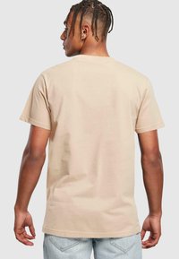 Beige bomulds t-shirt med korte ærmer, rund hals og løs pasform, vist fra ryggen mod en lys grå baggrund.