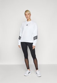 Hvit genser med svart Adidas-logo, svarte leggings med mesh-paneler, og hvite sneakers med svarte striper.