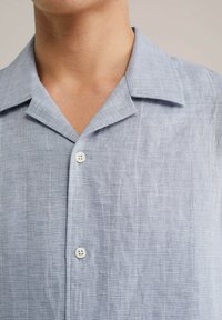 Camicia azzurra a maniche corte con un motivo testurizzato, colletto a punta e bottoni bianchi. Il tessuto sembra traspirante e morbido.