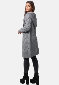 Cardigan grigio lavorato a maglia con cappuccio, caratterizzato da motivi a treccia, polsini e orlo a costine. Indossato sopra jeans skinny neri e stivali con tacco.