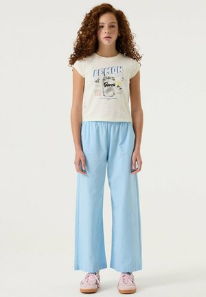 Mädchen mit lockigem roten Haar, das ein weißes T-Shirt mit Limonaden-Grafik, hellblaue Schlaghosen und pinke Sneakers trägt, steht vor einem einfarbigen Hintergrund.