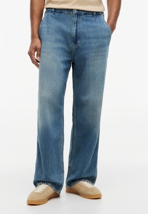 Man draagt lichtblauwe wijd uitlopende jeans en beige sneakers, staat met handen in de zakken tegen een effen achtergrond.