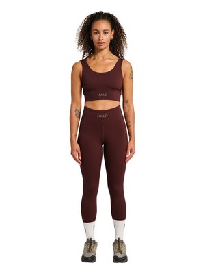 Kvinde står, iført matchende mørkebrunt sports-bh og høj taljeleggings med "HALO" logo, hvide sokker og vandresko.