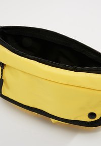 Keltainen kangaspussi, jossa on musta vetskari ja reuna, ja joka on varustettu sileällä pinnalla sekä pienellä mustalla nappikiinnityksellä.