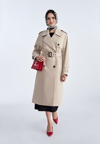 Beżowy trencz z podwójnymi guzikami, paskiem w talii i pagonami na ramionach. Noszony z czarną sukienką, czerwonymi szpilkami i wzorzystą chustą.