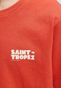 Sudadera de algodón roja con un gráfico de texto blanco que dice "SAINT-TROPEZ FRENCH RIVIERA". Cuello acanalado, textura suave.