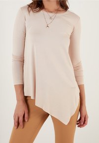 Top lungo rosa chiaro a maniche lunghe realizzato in tessuto morbido, con orlo asimmetrico, scollo rotondo e vestibilità slim. Indossato sopra pantaloni color sabbia.