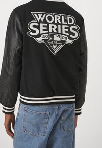 Person som bär en svart varsityjacka med lädersleeves och stor vit "World Series 2009 Fall Classic"-logotyp på ryggen, tillsammans med blåjeans.