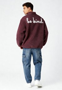 Giacca in pile color marrone con collo alto e testo bianco "be kind." sul retro. Abbinata a jeans cargo azzurri e scarpe da ginnastica bianche.