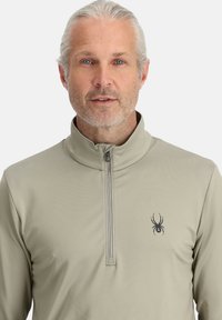 Licht beige quarter-zip pullover met een zachte textuur en een klein zwart spinnetje logo op de linkerborst. Uitgerust met een opstaande kraag.
