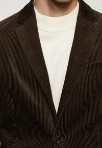 Mężczyzna w ciemnoszarym żakiecie z kordurowej tkaniny, z białym swetrem z okrągłym dekoltem, widoczna klatka piersiowa i szyja, bez widocznych cech twarzy.