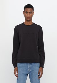 Black sweatshirt features a round neck and embossed "MICHAEL KORS" logo on the front. Made from a smooth fabric, paired with light blue denim jeans.