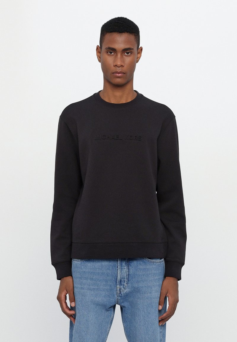 Black sweatshirt features a round neck and embossed "MICHAEL KORS" logo on the front. Made from a smooth fabric, paired with light blue denim jeans.