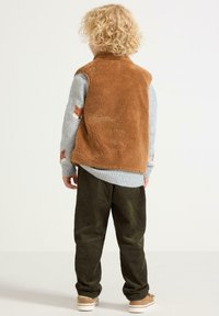Dziecko z kręconymi blond włosami, ubranym w brązową kamizelkę polarową, szary sweter z wzorami lisów, oliwkowe spodnie z kordurowej tkaniny i beżowe trampki, odwrócone plecami.