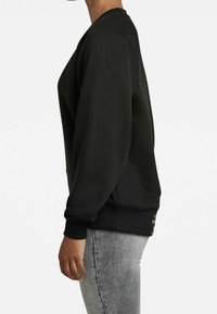 Sudadera negra con cuello redondo, puños acanalados y dobladillo. Presenta detalles laterales cerca del dobladillo. Combinada con jeans gris claro.