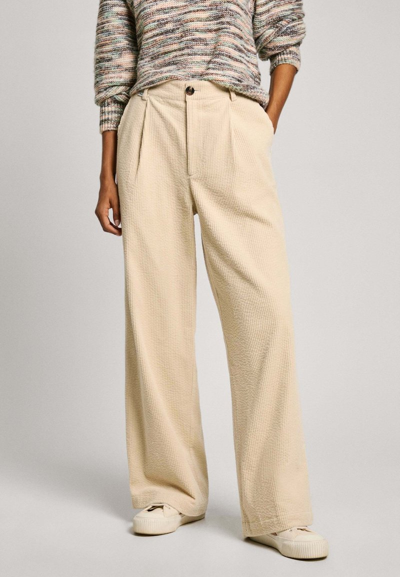 Personne portant un pantalon en velours côtelé beige à jambes larges avec fermeture à bouton et zip, associé à un pull tricot multicolore et des baskets crème.