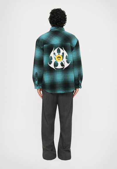 Camicia a quadri verde e nera con una grande grafica bianca e un volto giallo sul retro; maniche lunghe, polsini con bottoni, vestibilità rilassata.