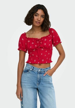 Femme aux cheveux ondulés et foncés portant un chemisier court rouge à fleurs et un jean bleu taille haute, une main dans la poche, sur fond blanc.