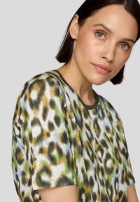 Frau mit kurzem dunklem Haar und heller Haut, die ein kurzärmliges, mehrfarbiges Leopardenmusterhemd vor einem schlichten hellgrauen Hintergrund trägt.