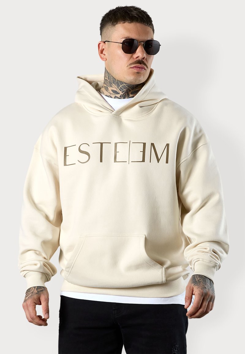 Sweat à capuche couleur crème avec grande poche avant, poignets côtelés et graphique "ESTEEM" en marron sur la poitrine. Matière en coton, coupe décontractée.