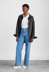 Black leather oversized shirt jacket, white striped polo, blue wide-leg jeans, and light pink sneakers. Smooth texture and casual fit.