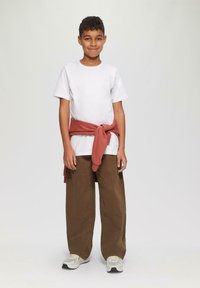 Jongen staat en glimlacht, draagt een wit T-shirt, bruine broek, witte sneakers en een rode sweatshirt om de taille geknoopt.