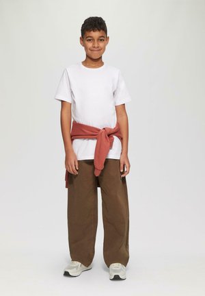 Jongen staat en glimlacht, draagt een wit T-shirt, bruine broek, witte sneakers en een rode sweatshirt om de taille geknoopt.