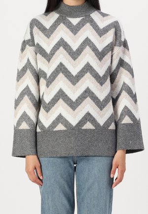 Frau trägt einen grau-weißen und beigen Chevron-Strickpullover mit breiten Bündchen und blaue Jeans, gezeigt vom Hals bis zur Hüfte.