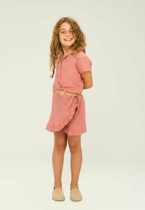 Pink two-piece outfit featuring a button-up top with a tied front, ruffled hem skirt, and beige slip-on shoes. Soft fabric texture.