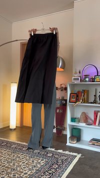 Black, knee-length skirt with an elastic waistband, held by a hanger, paired with gray, flared trousers, on display in an indoor setting.