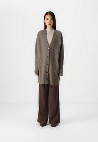 Cardigan oversized, maro, cu guler în V, cu șase butoane pe față, manșete cu textură dublă și o textură moale, pufoasă, asortat cu pantaloni maronii cu picioare largi.
