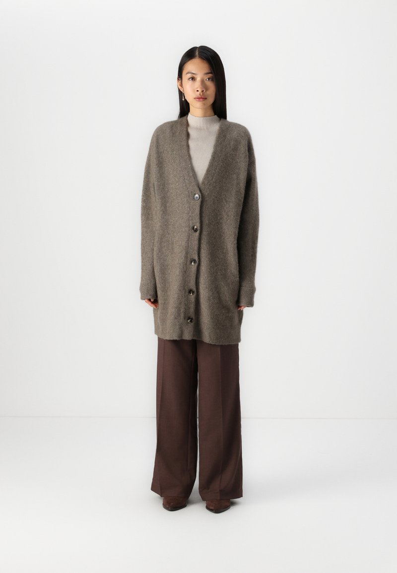Cardigan oversized, maro, cu guler în V, cu șase butoane pe față, manșete cu textură dublă și o textură moale, pufoasă, asortat cu pantaloni maronii cu picioare largi.