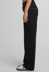 Black wide-leg trousers made of cotton, featuring a button at the waistband, side pockets, and worn with white and gray sneakers.