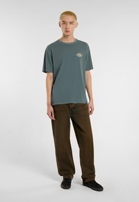 Camiseta de algodón verde grisáceo con mangas cortas y cuello redondo, que presenta un pequeño logo. Combinada con unos jeans de mezclilla marrones, holgados, y zapatos negros.