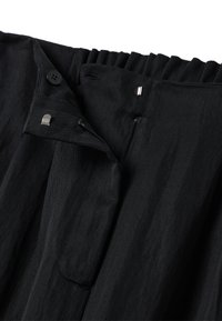 Tessuto nero con texture a righe verticali; presenta una chiusura con bottone e zip, e una tasca. Dettaglio della vita arricciata nella parte superiore.