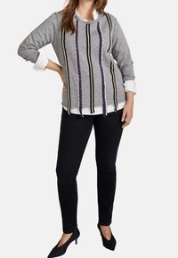 Grauer Pullover mit vertikalen Streifen aus schwarzem, beigem und lila Garn. Kombiniert mit schwarzen Hosen und schwarzen High Heels.