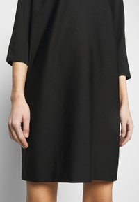 Femme portant une robe noire jusqu'aux genoux avec des manches trois-quarts, debout devant un fond clair uni.