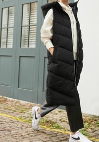 Czarna pikowana kamizelka z kapturem, z zamkiem z przodu i bocznymi kieszeniami, noszona na białej bluzie z kapturem, w połączeniu z czarnymi skórzanymi spodniami i białymi sneakersami.