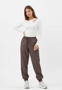 Hvid langærmet top med sideskæringer, parret med løse brune bukser med elastisk talje og manchetter ved anklerne. Grå sneakers.