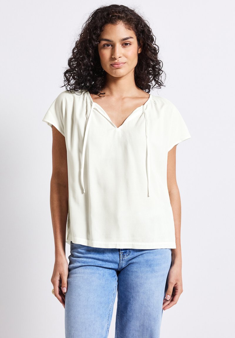 Joven con cabello rizado y oscuro, lleva una blusa blanca de manga corta y suelta, junto con unos jeans de color azul claro, de pie contra un fondo liso.