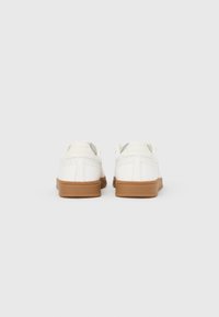 Ζευγάρι λευκών παπουτσιών χαμηλού τύπου με καφέ λαστιχένιες σόλες, εμφανιζόμενα από πίσω σε απλό λευκό φόντο.