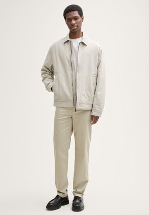 Veste légère beige avec fermeture éclair à l'avant, deux poches latérales, associée à un pantalon beige clair et des chaussures noires, sur un fond neutre.