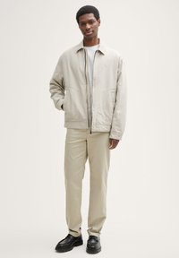 Jachetă bej deschis cu închidere cu fermoar, două buzunare laterale și guler ridicat, asortată cu pantaloni bej și pantofi negri.