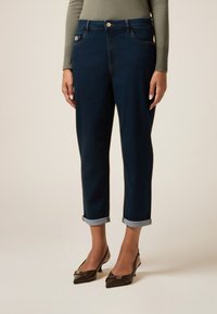 Jeans azul oscuro con una cintura ajustada, puños doblados, presillas para cinturón y un botón metálico en la parte frontal. Llevados con tacones marrones de punta.