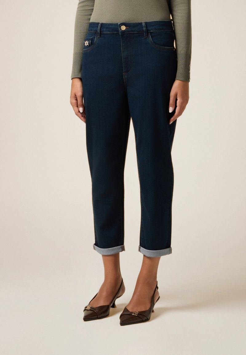 Jeans azul oscuro con una cintura ajustada, puños doblados, presillas para cinturón y un botón metálico en la parte frontal. Llevados con tacones marrones de punta.