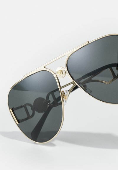 Ochelari de soare aviator aurii cu lentile întunecate, având un logo proeminent Medusa, ramă metalică și un design distinct al templelor.