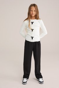 Vit långärmad topp med svarta rosett detaljer, volang nederkant, kombinerad med svarta vida byxor och svart-vita sneakers.